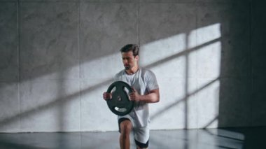 Güçlü sporcu hafif stüdyoda ağırlık plakası tutarak hamleler yapıyor. Spor eğitmeni. Çatı katındaki spor salonunda güç ve teknik gösterisi yapıyor. Egzersiz yapan motive bir adam.