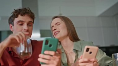 Akıllı telefonları olan aile çifti mutfaktaki masada kahve içerek gülüyor. Mutlu eşler cep telefonlarıyla ev kahvaltısının keyfini çıkarıyorlar. Neşeli gençler mobil teknolojiyle etkileşim halindeler.