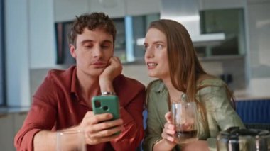 Akıllı telefon içeriğini tartışan erkek kadın daire mutfağında yakın plan çekimde oturuyor. Evde kahvaltıda kahve içen rahat bir çift. Mutlu eşler sabah etkileşiminden zevk alıyorlar