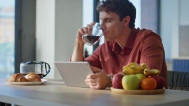 Freelancer mutfak masasında kahve görünümlü tablet ekranı içiyor. Evde rahatlamış bir adam sabah içeceğinin tadını çıkarıyor. İçerideki dijital cihazda sosyal medyayı izleyen sakin adam.