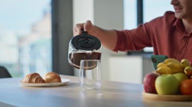 Rahatlamış bir adam kahvaltıda elinde akıllı telefon olan taze demlenmiş kahveyle bardağı dolduruyor. Sakinleştirici modern mutfakta cam bardağa espresso dolduruyor. Yakışıklı adam sabah içkisini yudumluyor..