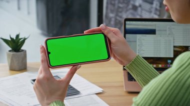 Modern ev ofisinde yeşil ekran akıllı telefon videosu izleyen kimliği belirsiz bir kadın. Kadın elleri yatay krom anahtar cep telefonunu tutuyor. Model cep telefonunda sosyal medya içeriği arayan Freelancer