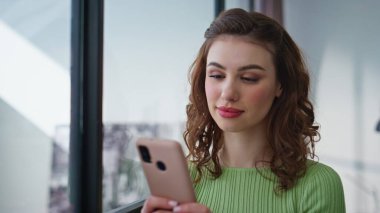 Gülümseyen bir çalışan modern panoramik ofiste akıllı telefon yazıları yazıyor. Duygusal kurumsal yönetici kadın barışçıl bir mola veriyor cep telefonu uyarısı alıyor.