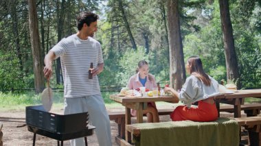 Aile, Sunny Park 'ta yaz barbekü pikniğinin keyfini çıkarıyor. İki kadın yeşil ağaçlarla çevrili masada otururken adam yemek pişiriyor. Gençler Bbbq toplantısını yiyecek içecekleriyle ve dostça konuşmalarla yapıyorlar.