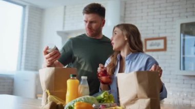 Mutfakta yemek sıralayan yakın bir çift. Aile, kağıt torbalardan satın alınmış ürünleri alıyor. Mutlu çift sağlıklı sebzelerle birlikte ekolojik süpermarket paketleri açıyor.