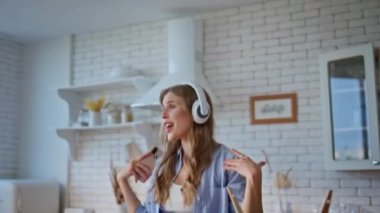 Modern mutfakta dans eden kulaklıklı kadın. Sağlıklı yemek yapan mutlu kız, çağdaş evde elinde akıllı telefonuyla sevinç çığlıkları atıyor. Kablosuz kulaklıklarla gülümseyen kadın, tek başına mutfakta. 