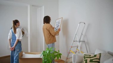 Kız arkadaş, erkek arkadaşının oturma odasını dekore edişini izliyor duvara resim asıyor. Kadın, kocasının modern resim için yer seçmesine yardım ediyor. Minimalist bir tarz yaratıyor. Genç aile yeni evinde rahat eder..