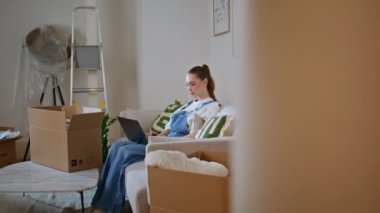 Kız alışveriş laptopunu rahat kanepe yeni kaydırak manzaralı internete yüklemek. Taşındıktan sonra bilgisayardaki e-ticaret mobilyalarına göz atan rahat genç bir kadın. Sakin bayan kutuların yanında oturuyor.