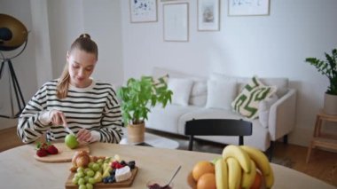 Parlak modern mutfakta elma doğrayan sakin bir kadın. Rahatlamış bir bayan kahvaltıda taze vitamin hazırlıyor. Gülümseyen at kuyruklu kız yeşil meyve kesiyor ve hafta sonunun tadını çıkarıyor..