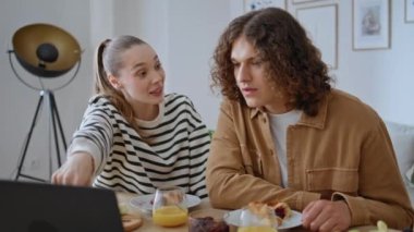 Birkaç endişeli dizüstü bilgisayar haberi modern mutfakta yakın plan kahvaltıda. Duygusal bir kadın dikkatle dinleyen bir adama bilgisayar ekranını işaret ediyor. Ciddi eşler dijital aygıt evinde bir şey tartışıyor.