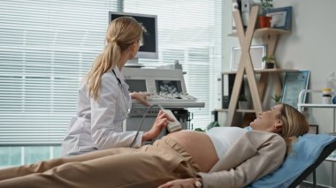 Klinikteki ultrason ziyaretiniz kutlu olsun. Gülümseyen doktor hamilelik taramasını monitörde gösteriyor. Muayene odasında müstakbel annesiyle konuşuyor. Kadın profesyonel sonogram makinesi kullanıyor.