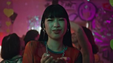 Neon parti atmosferinde konfeti üfleyen gülümseyen kadın. Gece kulübünde neşeli bir şekilde dans eden mutlu Asyalı kız. Parlak makyajlı, neşeli kadın milenyumu. Canlı diskoteğin tadını çıkarıyor..