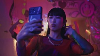 Kulüp partisinin neon ışıkları altında akıllı telefondan selfie çeken kulüpten bir kadın. Asyalı kadın blogcunun canlı gece hayatında cep telefonunda yüz fotoğrafı çekmesi. Sosyetik kız içerik sosyal medya yapıyor