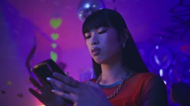Neon gece kulübü ışıklarında cep telefonuna göz atan bir kadın. Parlak diskotek atmosferinde modayı takip eden Koreli akıllı telefon. Modern kulüp tarzı ve sosyal etkileşim.