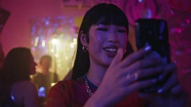 Neon ışıklı kulüpteki partide gülümseyen bir kadın. Neşeli bir gece kulübünde akıllı telefonda gece hayatını filme alan mutlu Koreli kız. Parlak makyajlı neşeli kulüp üyesi barda cep telefonu kamerasıyla konuşuyor.