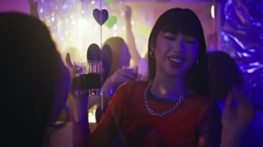 Gece kulübü partisinde elinde alkol bardağı tutan dansçı kadın. Mutlu Asyalı kız gece kulübünde kız arkadaşıyla içki tokuşturuyor. Kaygısız gençler neon ışıkta dinleniyorlar. Gece hayatının tadını çıkarıyorlar.