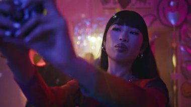 Parti kızı canlı neon gece hayatı kapanışında cep telefonuyla selfie çekiyor. Şık makyajlı Asyalı kadın etkileyicisi kutlama partisinde fotoğraf çekmenin tadını çıkarıyor. Neşeli kulüp üyesi yüz resmi çekiyor