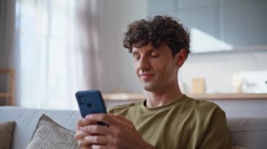Gülümseyen adam cep telefonunda SMS yazıyor rahat bir ev kanepesinde oturuyor. Neşeli yakışıklı adam dairede akıllı telefondan mesaj atıyor. Rahat erkek cep telefonu uygulaması rahat koltukta tek başına dinleniyor. 