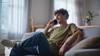 Oturma odasındaki kanepede cep telefonuyla konuşan gülümseyen adam. Dinlenen adam hafta sonu haberlerini akıllı telefonlardan aktarıyor. Mutlu hipster kanepede dinleniyor. Keyifli sohbetler. 