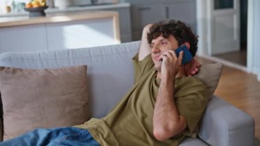 Haftasonu evde cep telefonuyla sohbet eden tembel öğrenci kendini rahat hissediyor. Pozitif erkek, elinde akıllı telefon ile kanepede konuşuyor. Yeşil tişörtlü gülen adam arkadaşça bir telefon konuşması yaptı.
