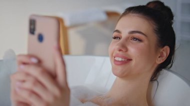 Gülümseyen kadın köpük banyosunda cep telefonuna mesaj atıyor. Neşeli esmer, akıllı telefonlu beyaz köpüklü banyo küvetinde rahatlamış. Banyoda dinlenen mutlu kız mesajlaşması..