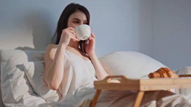 Güneşli bir otel odasında kahve içen bir bayanı aramak. Gülümseyen kadın akıllı telefondan konuşuyor rahat ev yatağında aromatik bir içecek yudumluyor. Mutlu kız yatak odasında cep telefonu sohbetinin keyfini çıkar..