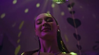 Neşeli kadın, mutlu bir gülümsemeyle diskotek ışıklarında hareket ediyor. Kaygısız kız neon ışıkta dans edip hafta sonu partisinin tadını çıkarıyor. Canlı disko topunun altında vücudunu hareket ettiren zarif bir kadın..