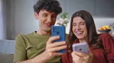 Gülen çift oturma odasında cep telefonu görüntülerine tepki veriyor. Mutlu sevgililer kanepede kaynaşıyor ve komik içeriğin tadını çıkarıyor. Kıvırcık çocuk ve gülümseyen kız akıllı telefonlar kullanarak yumuşak kanepeye bakıyor.