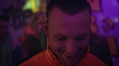 Neon ışıklı partide poz veren gülümseyen dövmeli adamın portresi. Karanlık dans pistinde dikilen neşeli kulüp üyesi parlak ışıklarla kameraya yakından bakıyor. Mutlu hipster parti kulübü atmosferinin tadını çıkarıyor..