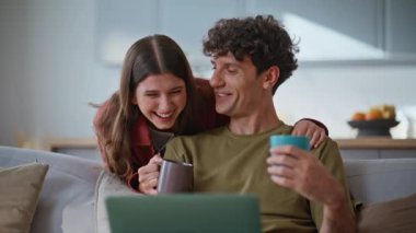 Modern dizüstü bilgisayarları kullanarak evde film izlerken gülen çiftler. Gülümseyen genç eşler kanepede rahat bir şekilde oturup kahve fincanı içerek komik filmlerin tadını çıkarıyorlar. Rahatlamış aile hafta sonu. Evlilik rutini 