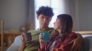 Kanepenin içinde çay fincanı tutan konuşan eşler. Aşk çifti evde kahve içip eğleniyor. Gülümseyen koca karısı sıcak içecek, rahatlatıcı kanepe içiyor. İnsanlar ev yaşam tarzı konsepti 