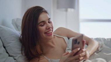 Sabahın erken saatlerinde yatağın üzerine akıllı telefon mesajı atan bir kız. Beyaz yatak yastıklarında uzanan neşeli kadın cep telefonu ekranına bakıyor. Neşeli esmer, güneşli yatak odasında çevrimiçi etkileşimden hoşlanıyor..