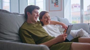 Tablet kullanan aşıkları kucaklamak. Yakın çekimde gülmek. Genç ve neşeli bir çift oturma odasında modern tablet bilgisayarlarını tutarak kanepeye sarılıyor. Pozitif aile eğlenceli videolar izlemekten hoşlanır.