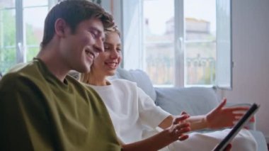 Mutlu çift, rahat ev çekimlerinde video çağrısı tableti yapıyor. Romantik gençler el sallayarak bilgisayar kameralarına bakıyorlar. İnternet üzerinden arkadaşlarıyla selamlaşan gülümseyen gençler 