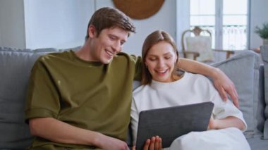 Modern evlerde tablet bilgisayarı arayan heyecanlı genç çift. Duygusal sevecen insanlar oturma odasındaki koltukta dijital cihaz kullanıyorlar. Mutlu gençler evde birlikte vakit geçiriyorlar. 