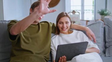 Kapalı çekimde konuşurken tablet kullanan sarılan çift. Kanepede kucaklaşan neşeli genç çift oturma odasında modern tablet bilgisayarlarını tutuyorlar. Pozitif aile hafta sonu evinde rahatlarken birlikte vakit geçirmeyi sever.