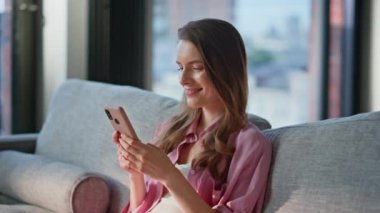 Rahat ev ortamında cep telefonu koltuğu kullanan gülümseyen bir kadın. Samimi oturma odasında iletişim akıllı telefonunun keyfini çıkaran mutlu esmer kız. Sakin bir bayan koltukta cep telefonu uygulamasına bakıyor.