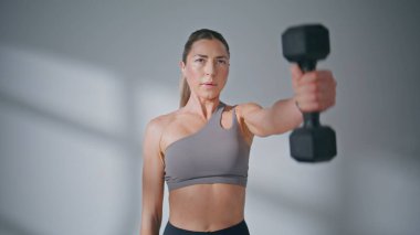 Sporcu kadın stüdyo kapanışında dambılları kaldırarak el sıkışıyor. Spor giyim sektöründe odaklanmış atletik bir kadın güneş ışığı odasında güçlü bir vücut için egzersiz yapıyor. Spor malzemeleriyle ciddi kız egzersizleri. 