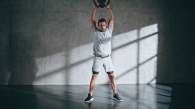 Çatı katında ağırlık çalışması yapan sporcu bir adam. Güçlü güçlü esmer, ağır ekipman kaldırıyor gri duvar arka planında kollarını kaldırıyor. Spor kulübünde tek başına omuz çalışması yapan ciddi bir sporcu. 