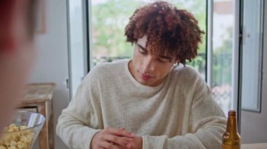 Latin teen enjoy boardgame at living room closeup. Curly millennial smiling to friends drinking beer during home party. Happy guy spending time with buddies at table with snacks. People lifestyle