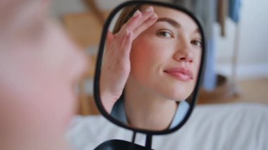 Gülümseyen kız yatak odasında tenine dokunuyor. Mutlu kadın sabah evde ayna yansıması görüyor. İçeride güneş ışığında sivilceleri kontrol eden güzel bir model. Cilt rutini kavramı 