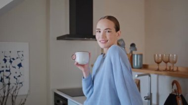 Uyanmış bir kız mutfakta cappuccino içiyor. Sabah yakın çekim sırasında sakinlik hissediyor. Kibar bir kadın elinde aromalı içecek bardağıyla dairede kameraya bakıyor. Ev elbiseli bayan günün tadını çıkarın.