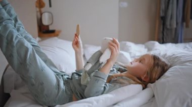 Yatak odasında ayna tutan küçük bir çocuk. Sakin bir hafta sonu sabahında pijamalı, yatakta uzanmış yansımaya bakan rahat bir çocuk. Küçük kız pazar gününün tadını çıkarıyor. Odanın içinde kaygısız hissediyor.