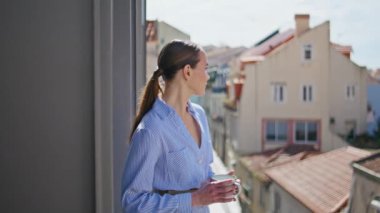 Güneşin altında elinde çay fincanı tutan romantik kız şehir manzaralı balkona yakın çekim yapıyor. Sabah terasında kahve fincanı taşıyan mutlu bir bayan model. Gülümseyen esmer kadın, kasabayı düşünüyor, yalnız başına dinleniyor.