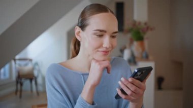 Rahatlamış bir kadın, sabahın erken saatlerinde mutfakta cep telefonuna bakıyor. Tasasız kız akıllı telefon ekranında evde oturuyor. Beyaz iç mekanda cep telefonu tutan sakin bir bayan ev huzurunun tadını çıkarıyor. 