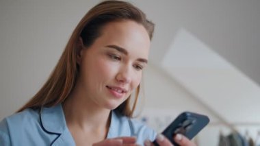 Pijamalı kız yatak odasında akıllı telefondan mesaj atıyor. Evde sakin bir sabah keyfi yapıyor. Rahat bir kadın cep telefonuyla yatağında sosyal medya izliyor. Sakin bayan internetten cep telefonu kullanıyor. 