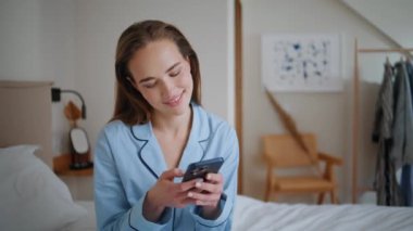 Hafta sonları cep telefonuna göz atan sabah kadını. Tasasız kız beyaz yatak odasında oturan akıllı telefon ekranına benziyor. Yatak odasında cep telefonu tutan rahat bayan huzurun tadını çıkarıyor. 