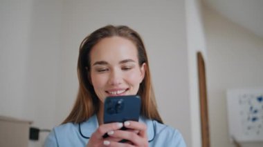 Gülümseyen bayan cep telefonuyla rahat bir şekilde yatak örtüsünde oturuyor. Kaygısız güzel kadın aydınlık yatak odasındaki akıllı telefondan sosyal medya içeriğini araştırıyor. Gün ışığında cep telefonuna mesaj atan mutlu kız. 