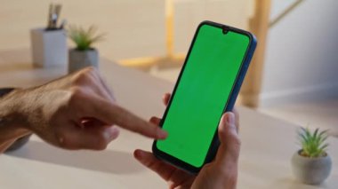 Chroma Key cep telefonundan alışveriş yapmayı seçen adam modern ev işyerinde yakın çekim yapıyor. Ofisteki yeşil ekranda akıllı telefon uygulaması kullanan bilinmeyen serbest çalışan. İşadamı elleri model cep telefonuna bağlanıyor