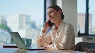 Gülümseyen iş kadını cep telefonunu arıyor. Ev ofisinin bilgisayarında çalışıyor. Yakın plan. Akıllı telefonla iş konuşmasına giren neşeli bir şirket çalışanı. Rahat kadın içeride cep telefonuyla konuşuyor.